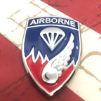 An Airborne military emblem badge featuring a shield design with a blue top half, red bottom half, white parachute and wing motifs, and the word AIRBORNE at the top, photographed close-up against a red-and-white background.
