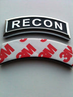 Two curved black and silver metallic emblem badges, one featuring the word 'RECON' in white and the other showing the red 3M logo, displayed on a neutral gray background.