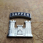 A silver metal enamel pin featuring a castle design and the word 'SAPPER' on a black arched tab, placed on top of an antique-style map background.